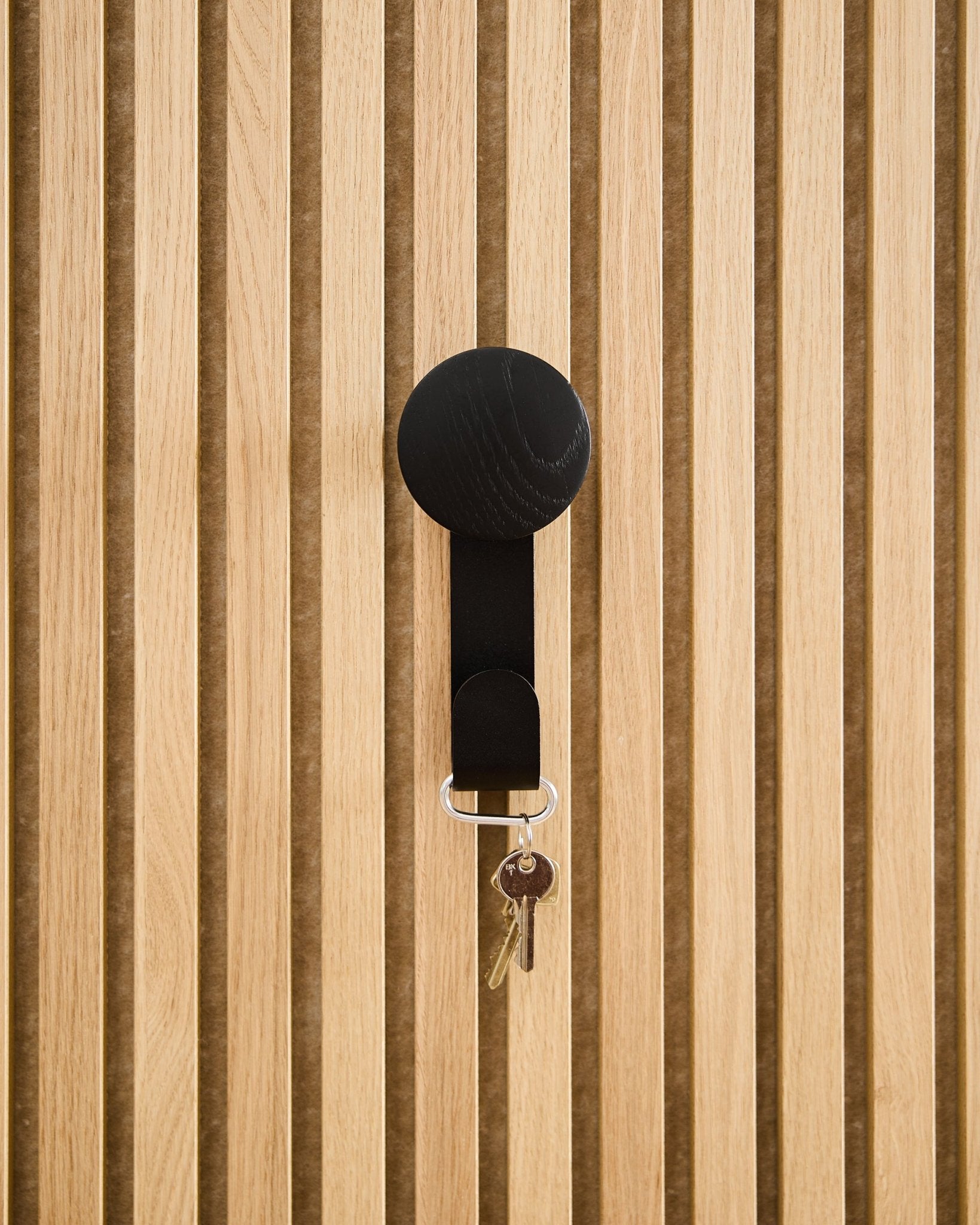 En Linn Vertical Hanger i Black Oak från WoodUpp håller en nyckelring med två nycklar och en rund bricka, monterad på en texturerad Akupanel akustikpanel i trä.